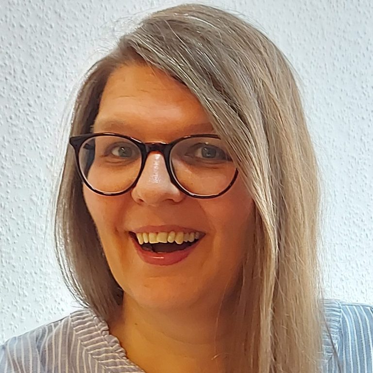 Lächelnde Frau mit langen blonden Haaren und Brille, weißer Hintergrund.